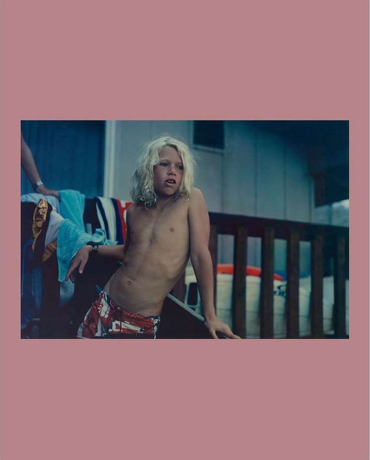 A shirtless boy with long blonde hair stands on a porch with towels and railings in the background on a sunny day.
