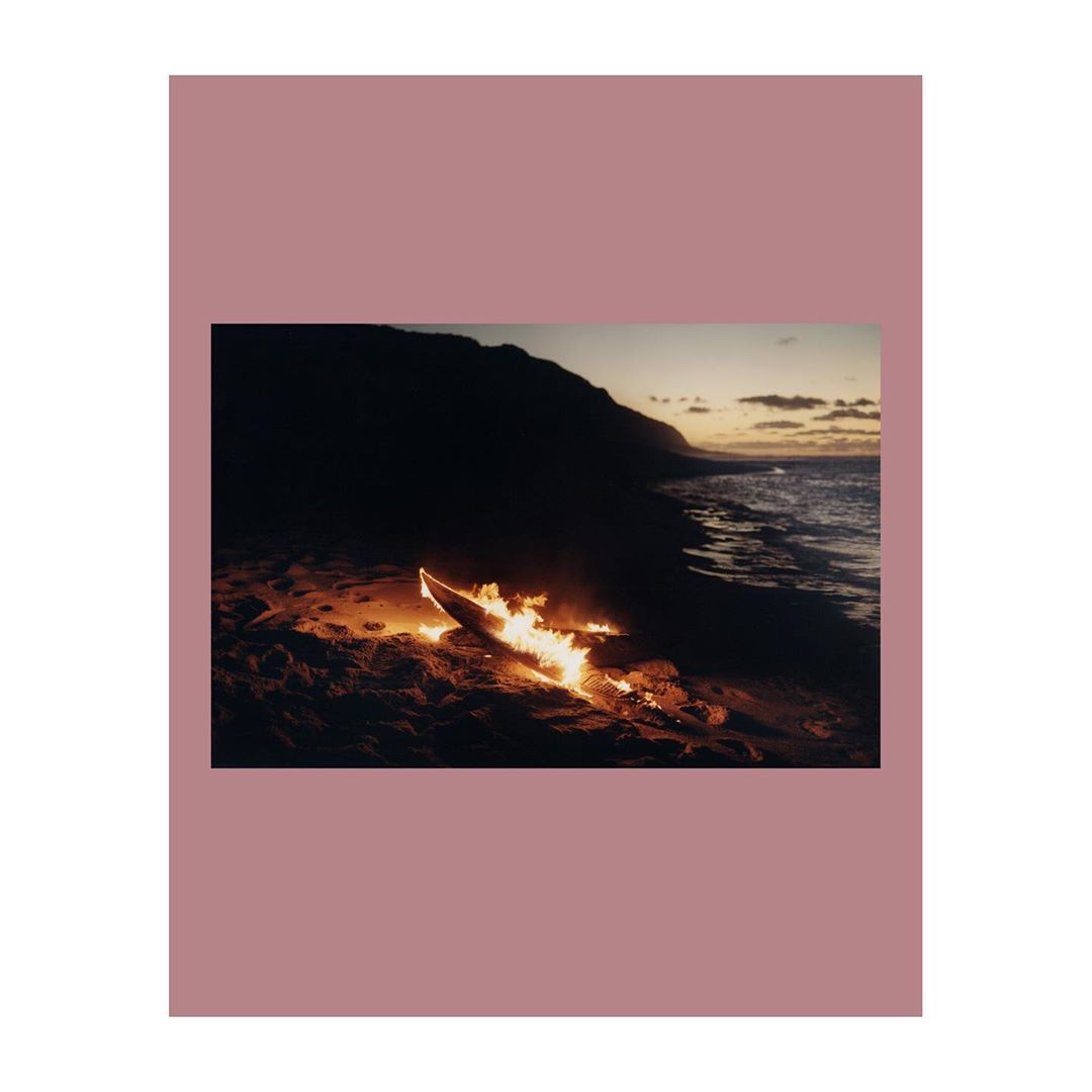 Surfboard on fire on a sandy beach, next to the ocean with mountains in the distance during dusk with a moody, muted color palette.