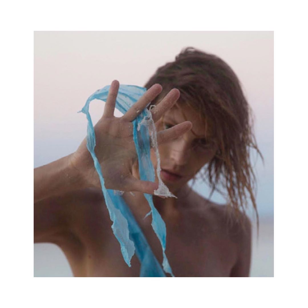 Portrait of a shirtless adult woman holding sheer blue fabric in front of her face at the beach with fair skin and wet brown hair.