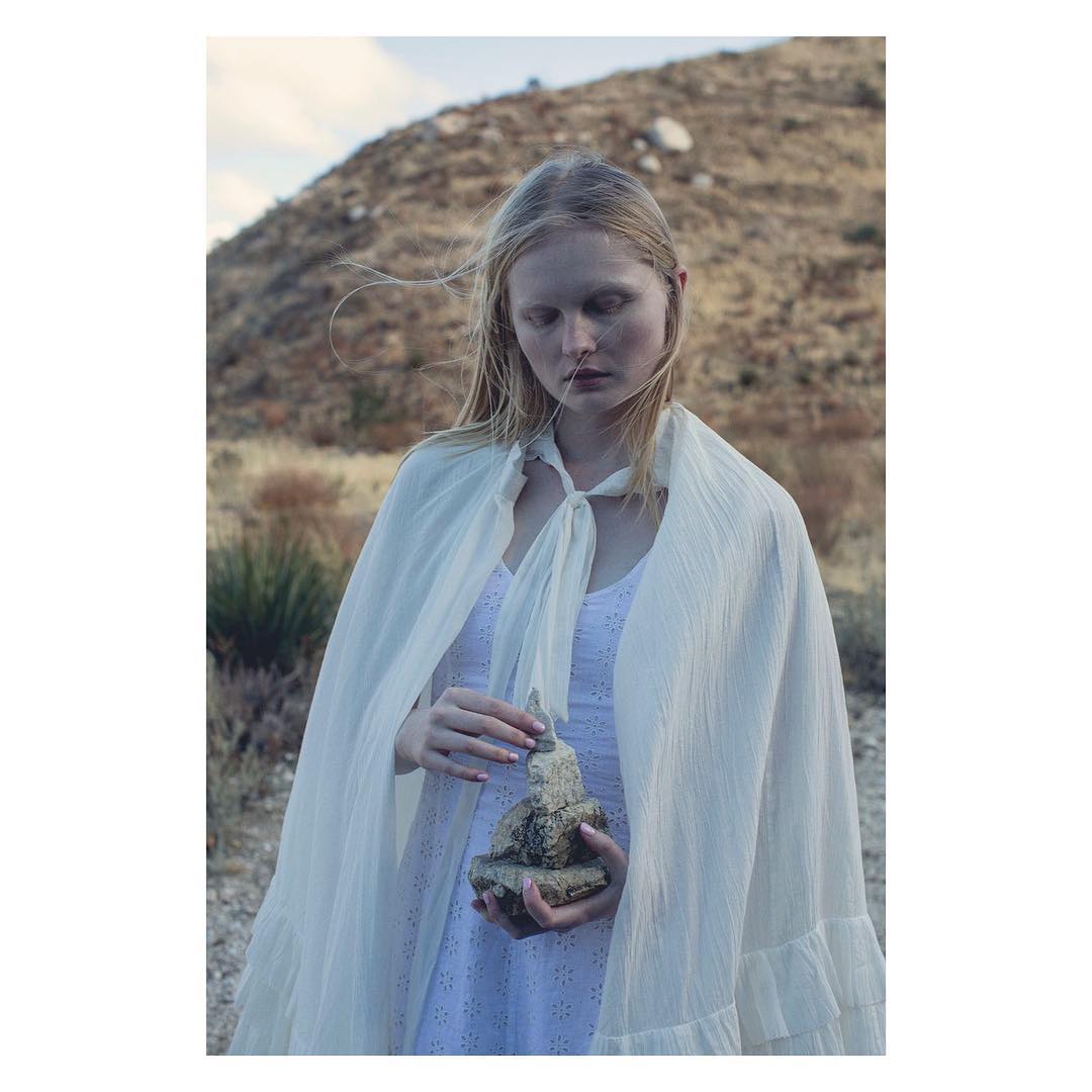 Woman in white dress and cape holds a small stone figure outdoors with a hillside backdrop in a vertical medium shot, evoking a peaceful mood.
