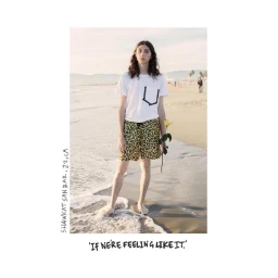 A long-haired young person in leopard print shorts and a white t-shirt holding a yellow flower standing on a sandy beach.