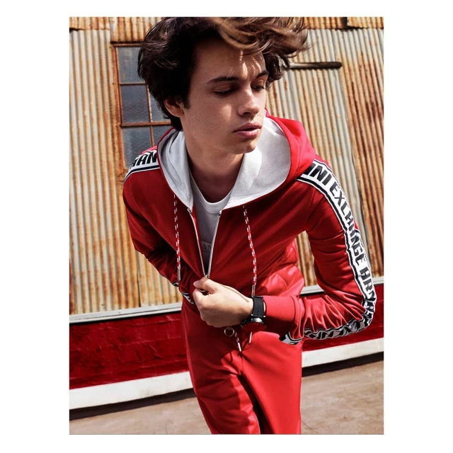 A young man in a red hooded tracksuit poses outdoors against a textured metal wall, showcasing a casual, athletic style and portrait framing.
