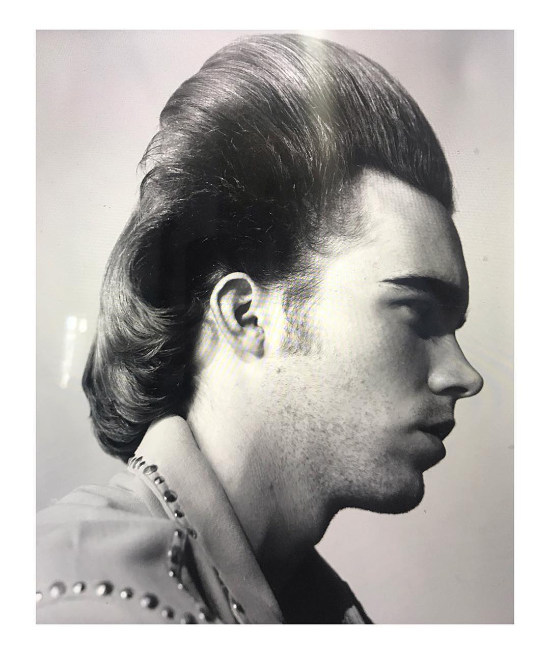 Profile portrait of an adult male with a vintage hairstyle, styled jacket, and a light background in a black and white photograph.