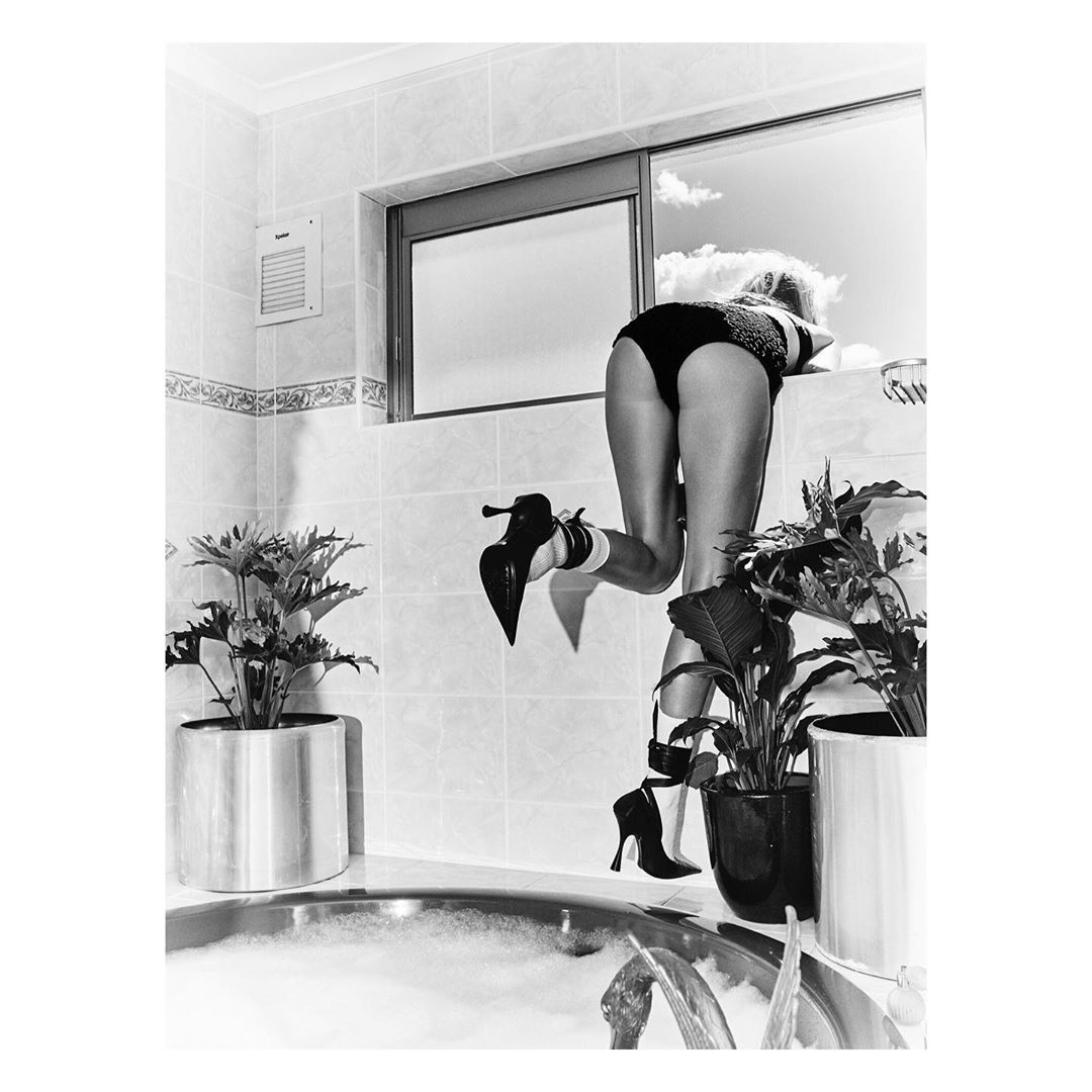 Black and white photograph of an adult female in a swimsuit and high heels posed in a bathroom with indoor plants and a view to the sky.