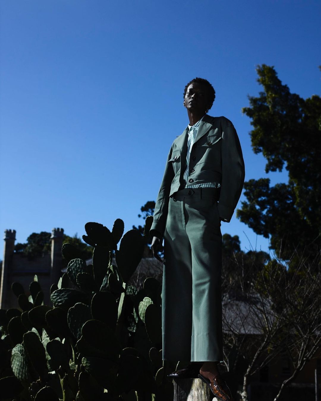A stylish African-American man in a green suit stands confidently in an outdoor setting, featuring cacti, clear blue sky, and old buildings.