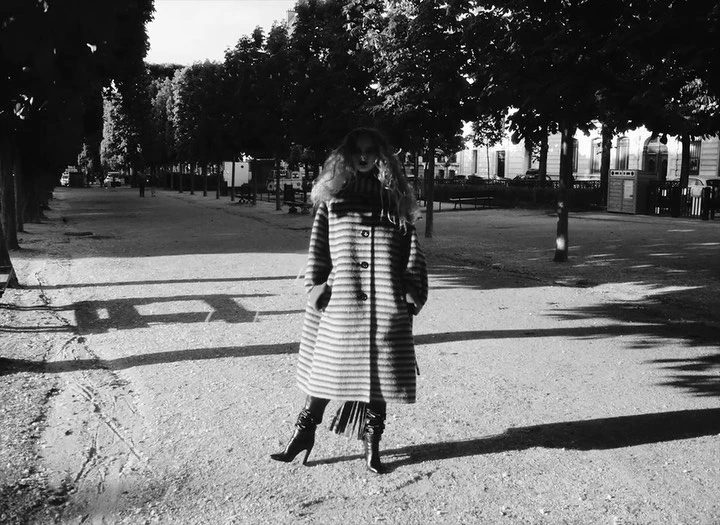 Woman in a striped coat and high-heeled boots stands in a park, the sun casting shadows on the ground, creating a monochromatic fashion shot.