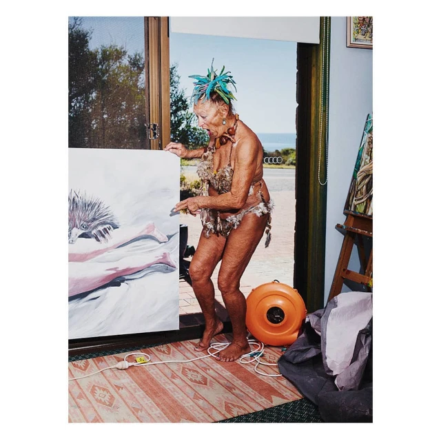 An elderly woman wearing a feather headpiece, standing next to a painting while holding a paintbrush inside a blue-walled building.