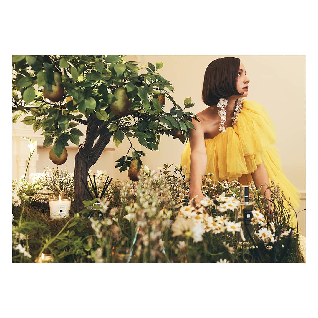 Woman in yellow tulle dress poses with pear tree, daisies, and perfumery products. Close-up in a dreamy indoor garden setting.