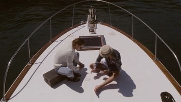 High-angle shot of two people seated on the bow of a white boat playing cards on the deck, surrounded by dark water