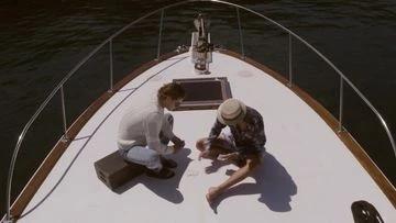 High-angle shot of two people seated on the bow of a white boat playing cards on the deck, surrounded by dark water
