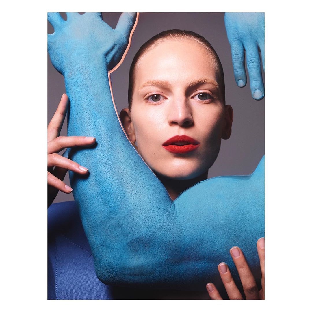 Close-up portrait of a woman with red lipstick framed by painted blue arms and hands against a grey background.
