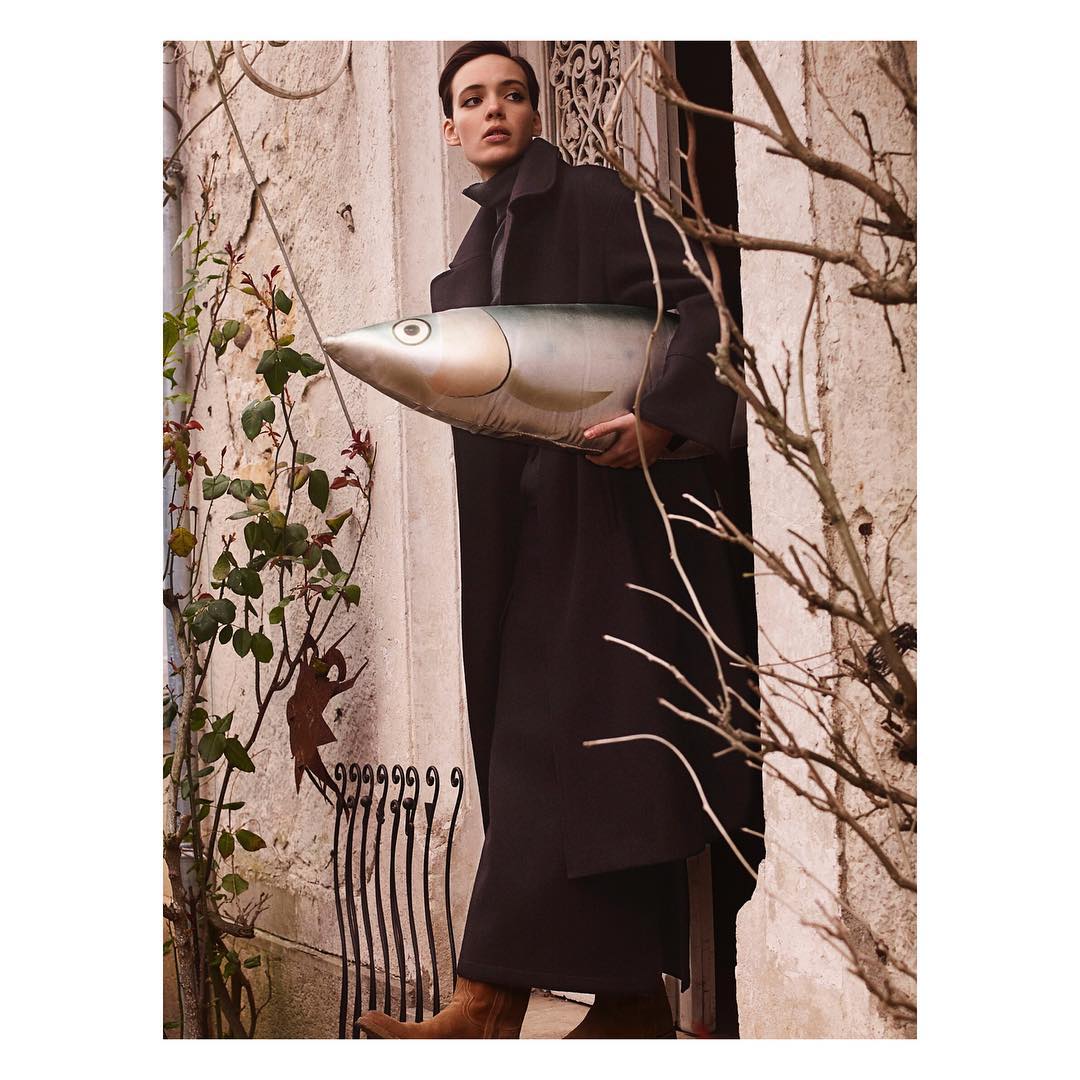 Adult woman in a black coat stands in a stone archway holding a large inflatable fish against a weathered stone wall with foliage.