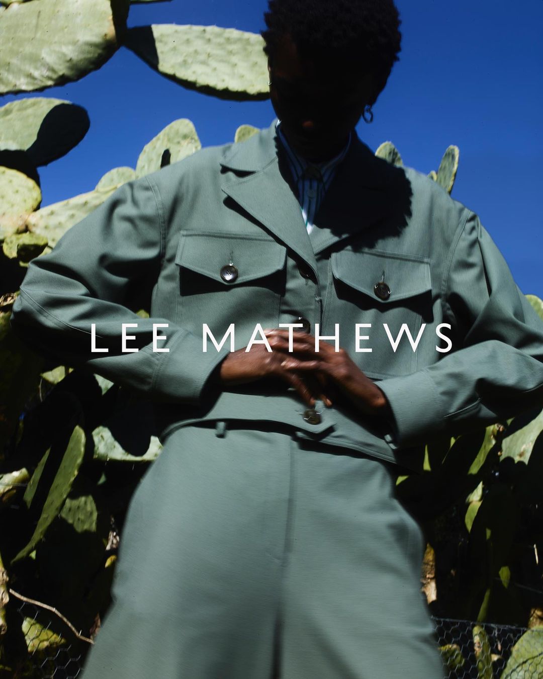 Model in sage green jacket and trousers posing against large cactus plants under a bright blue sky, photographed by Simon Eeles.