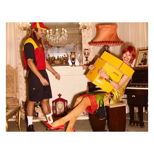 A woman sits holding DHL boxes in a stylized interior, while a man in a DHL uniform stands beside her in a colorful, theatrical image.