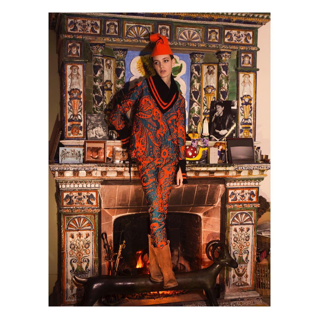 A woman in a paisley print suit and red hat stands in front of an elaborate fireplace with decorative tile and sculpture.