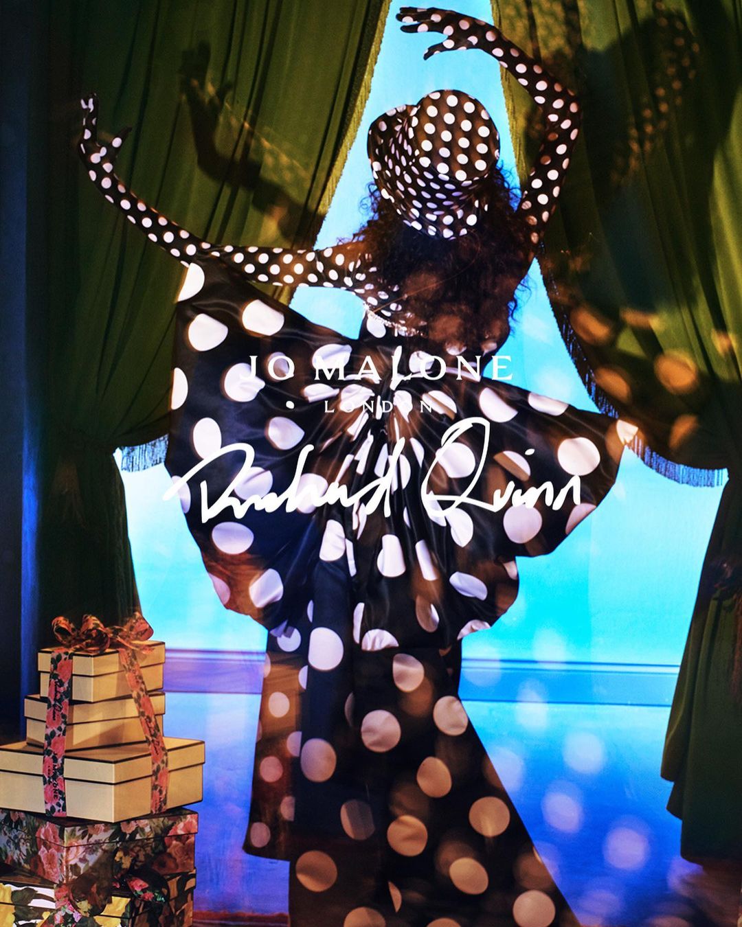 Woman in black and white polka dot dress strikes a pose with gift boxes in front of a window with green curtains and blue backdrop.