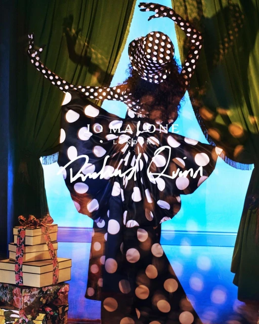 Woman in black and white polka dot dress strikes a pose with gift boxes in front of a window with green curtains and blue backdrop.