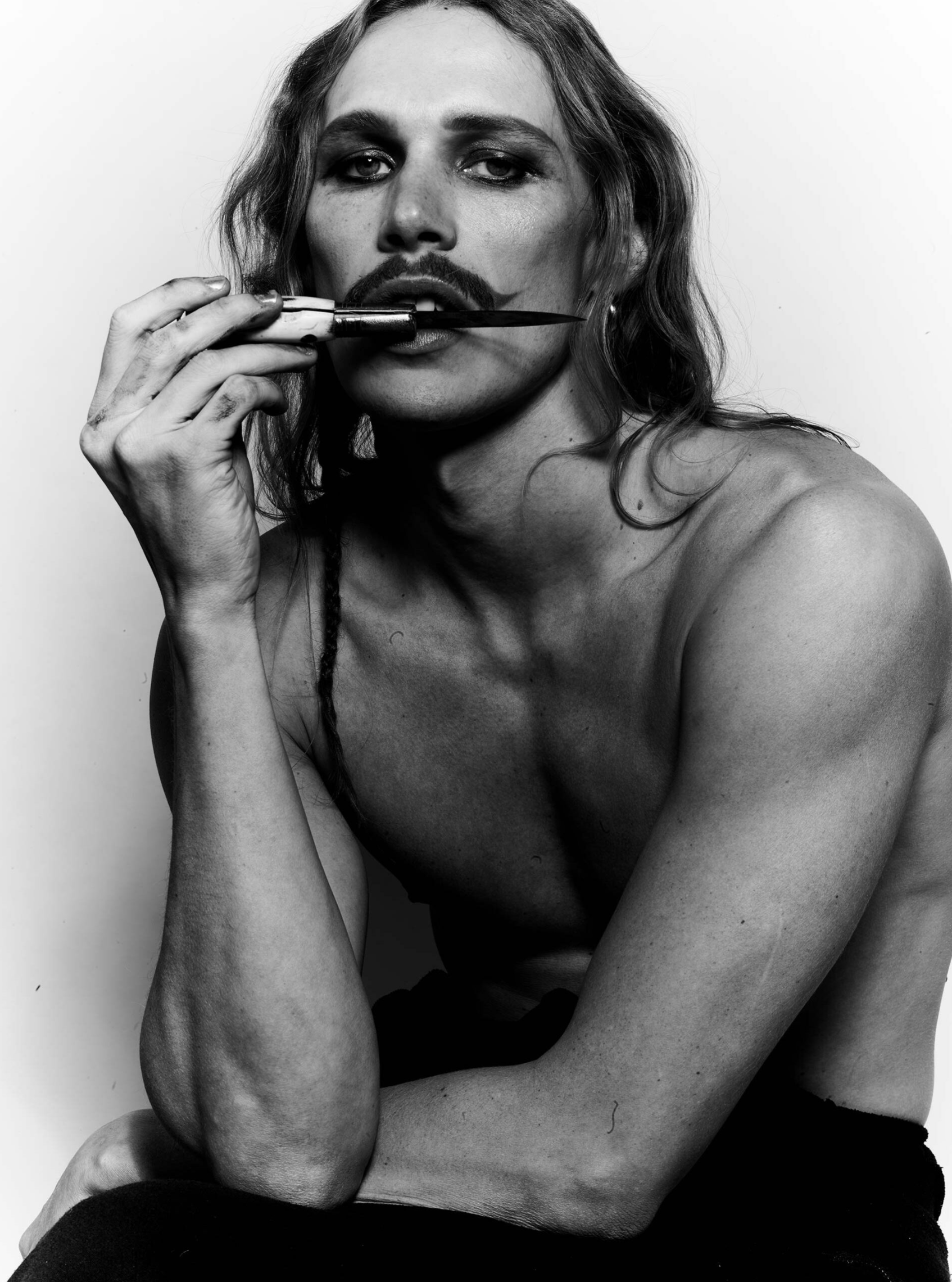 Striking black and white portrait of a person with long hair and a prominent moustache holding a knife to their mouth.
