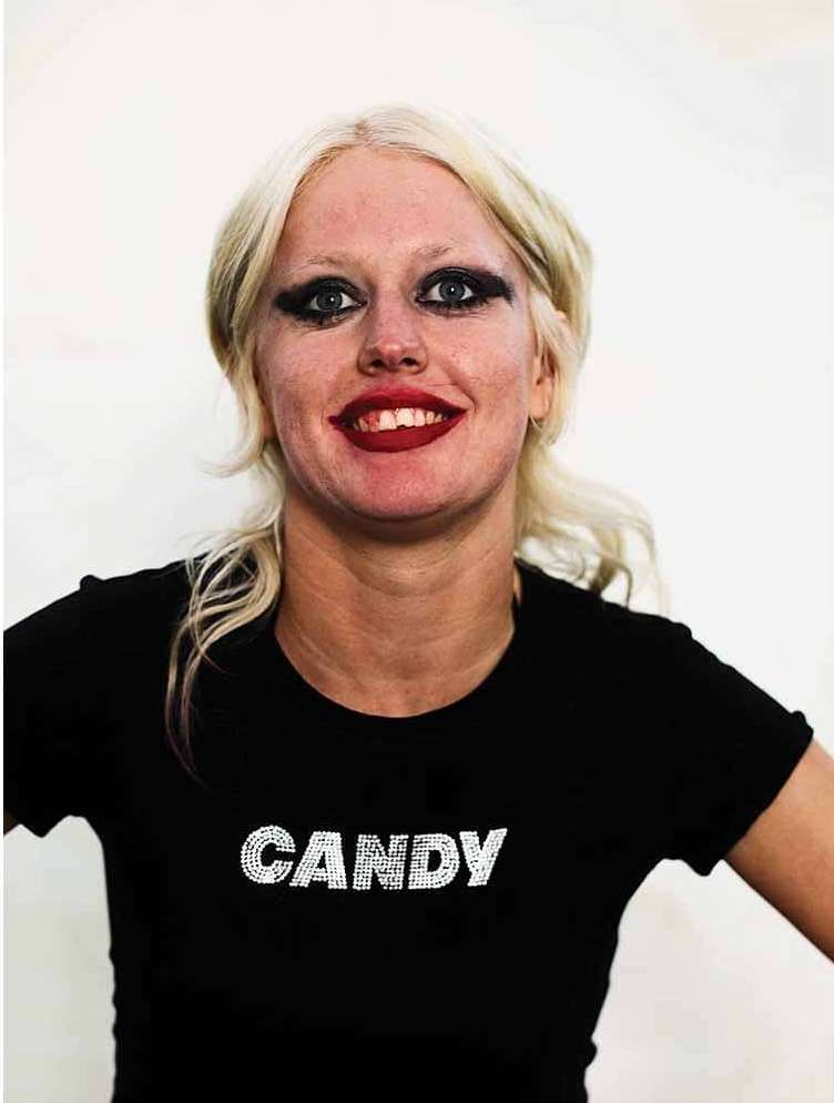 Portrait of a smiling adult woman with heavy makeup, blonde hair, and a black t-shirt with "Candy" written on it against a white backdrop.