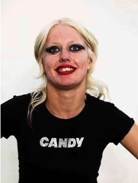 Portrait of a smiling adult woman with heavy makeup, blonde hair, and a black t-shirt with "Candy" written on it against a white backdrop.