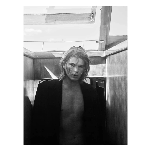 Monochrome portrait of a young adult male with long hair, wearing a dark blazer, posing indoors with neutral expression, and natural lighting.