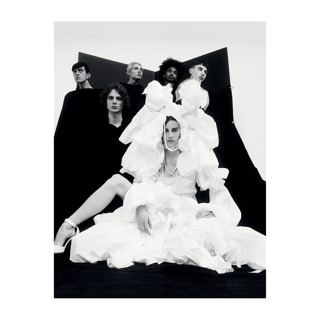 Group of six people wearing black and white clothing pose in a photo studio with white walls and black geometric shapes.
