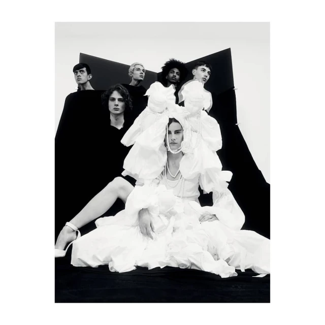 Group of six people wearing black and white clothing pose in a photo studio with white walls and black geometric shapes.