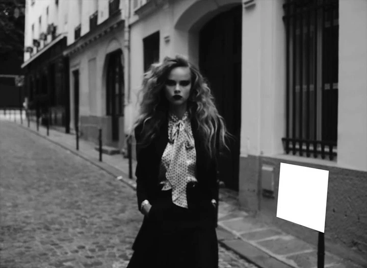 Elegant woman with long wavy hair stands on a cobblestone street in a black blazer, skirt, and a white polka dot blouse in an urban setting.