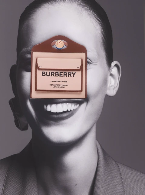 Burberry branded pouch with face illustration covering a model's face, black and white portrait background, fashion-forward shot.
