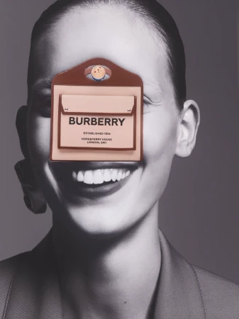 Burberry branded pouch with face illustration covering a model's face, black and white portrait background, fashion-forward shot.