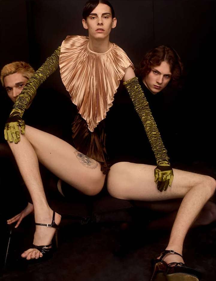 Three people wearing high fashion garments posed against a dark background, with the primary subject in a gold pleated collar.