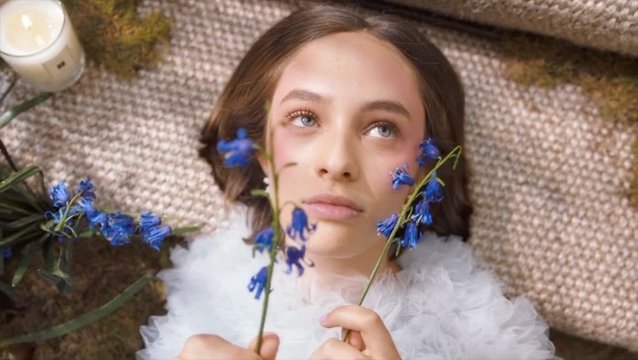 Close-up portrait of a woman laying on a textured surface holding bluebell flowers with a lit candle in the background.