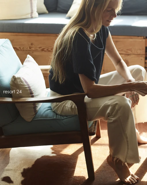 Adult woman sitting in a chair with pillows, wearing a navy shirt and white pants, indoors with a cowhide rug and wooden wall.