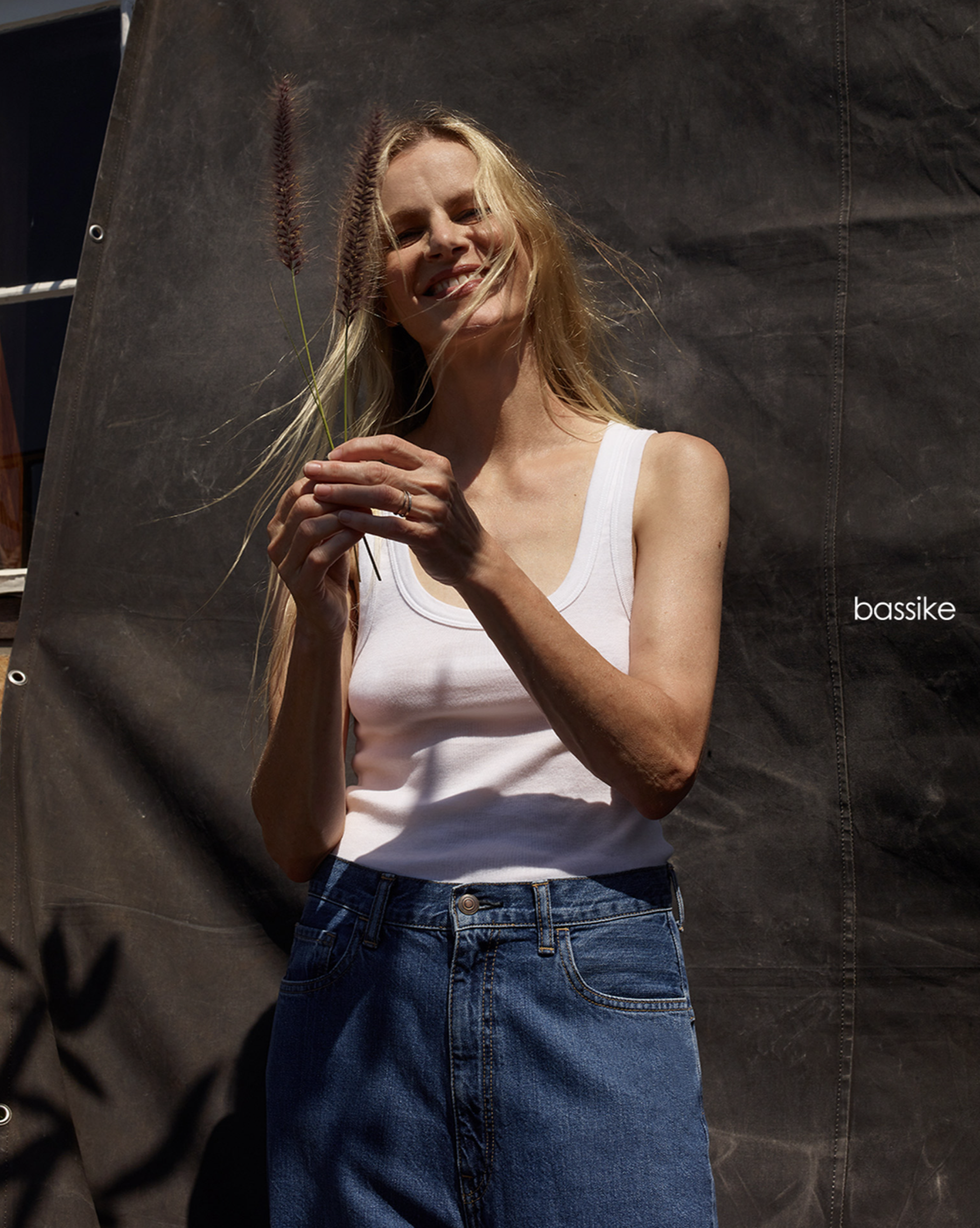 Smiling woman in a white tank top and blue jeans holding a plant in an outdoor setting against a dark fabric backdrop.