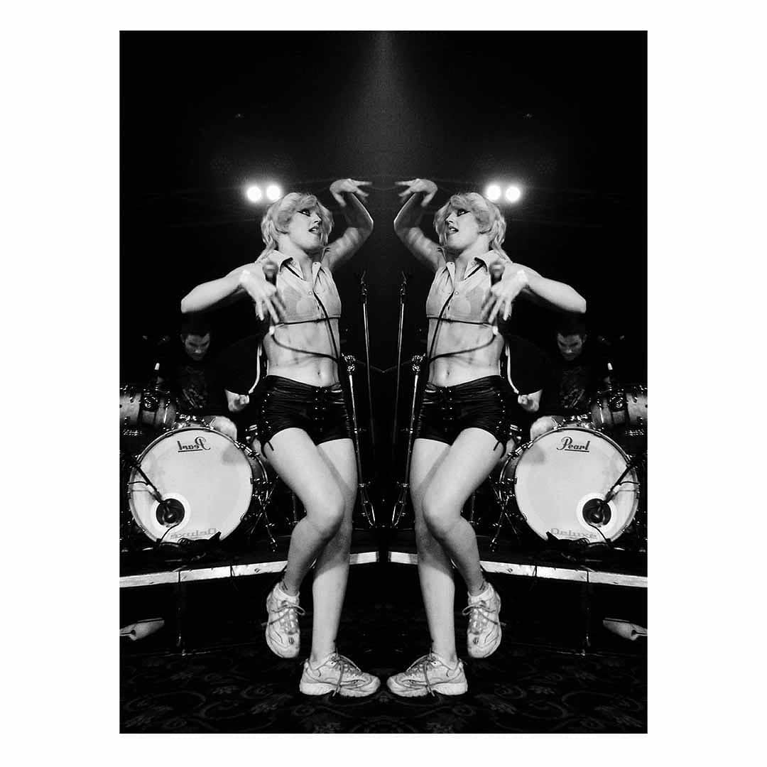 Mirrored black and white shot of a performer on stage in shorts and top dancing in front of drums, microphones, and band members.