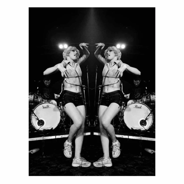 Mirrored black and white shot of a performer on stage in shorts and top dancing in front of drums, microphones, and band members.