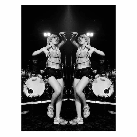 Mirrored black and white shot of a performer on stage in shorts and top dancing in front of drums, microphones, and band members.
