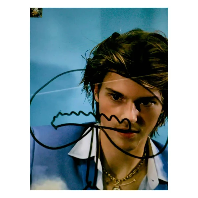Portrait of a young adult with brown hair, wearing a blue button-down shirt and a gold chain, in front of a light blue background.
