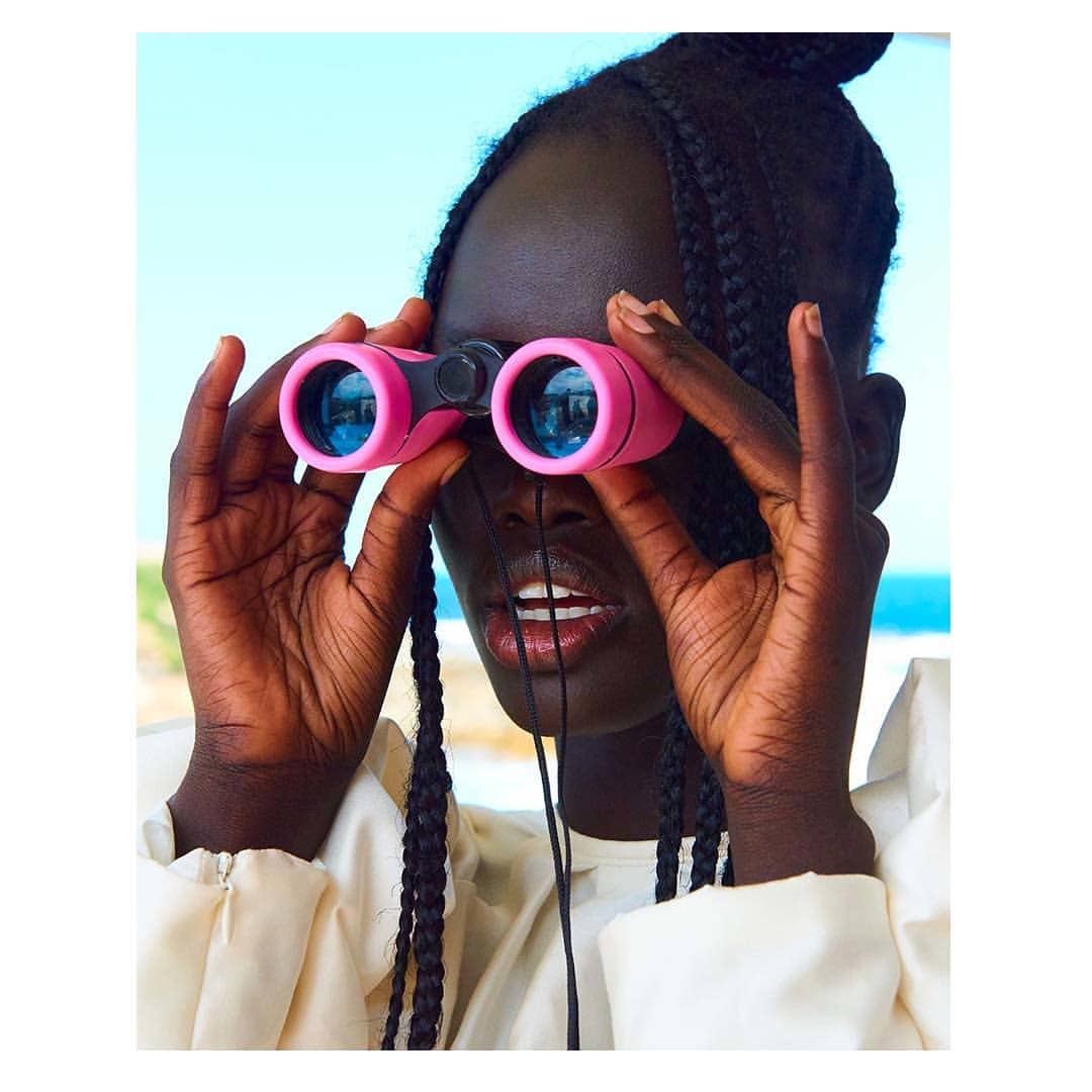 A woman with dark skin and braided hair is looking through pink binoculars against a bright blue sky in a close-up shot.
