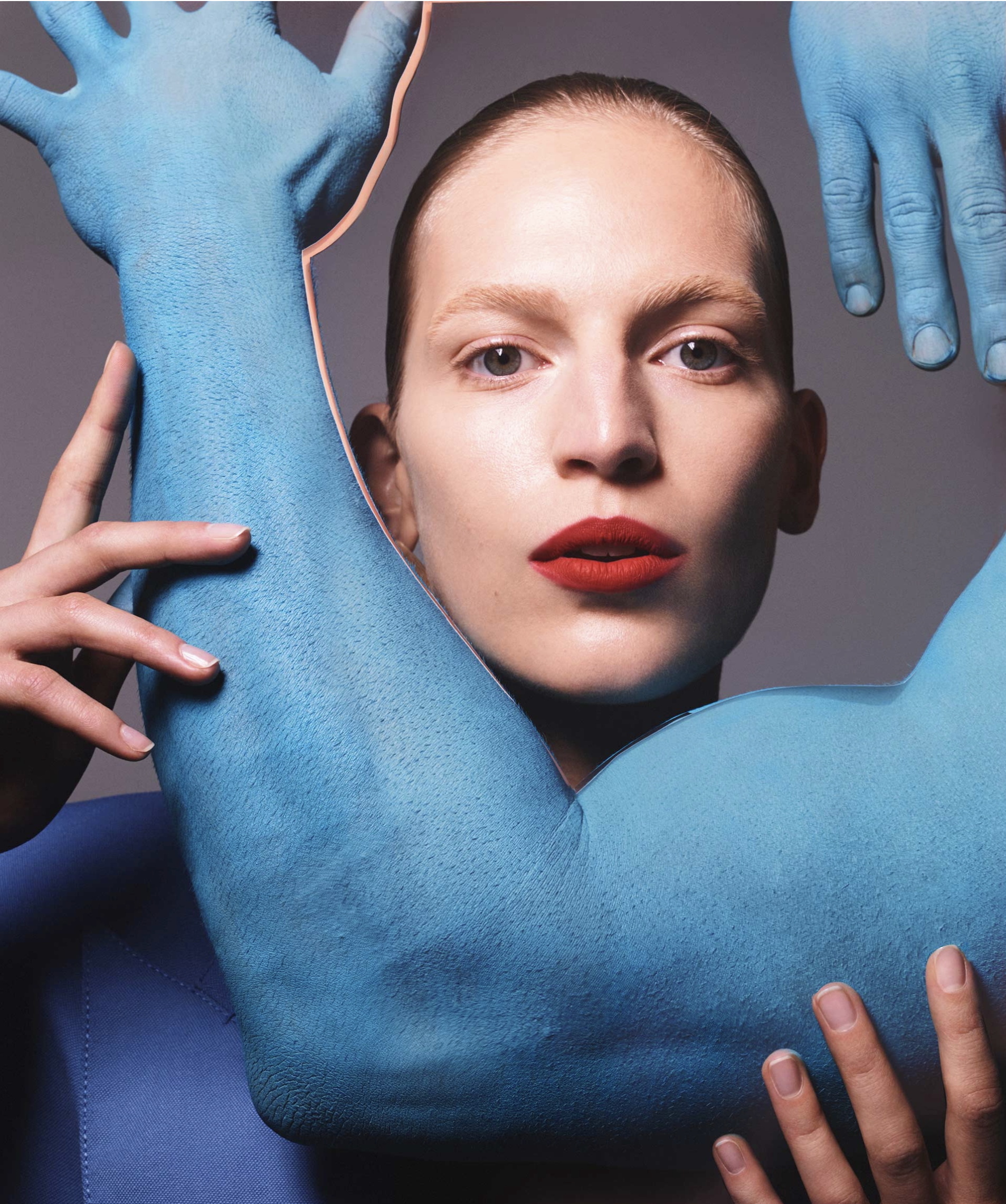 Woman with red lipstick poses with blue-painted arms and hands around her face against a grey background in a beauty-style portrait.