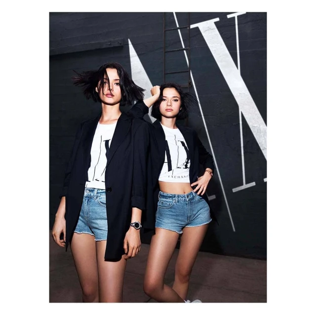 Two young women in casual fashion posing against a wall with the large AX logo. The photo shoot features urban style and modern design.