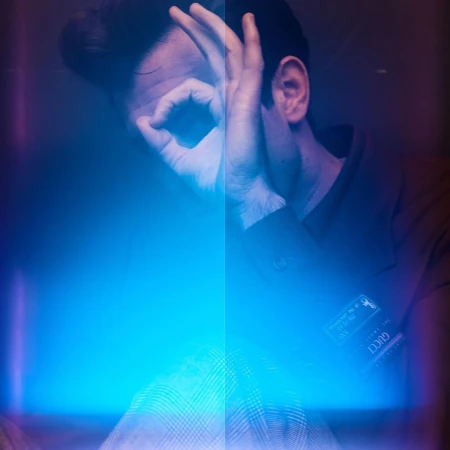 Close-up portrait of a man making an okay hand sign over his eye under dramatic blue and purple lighting by photographer Simon Eeles.