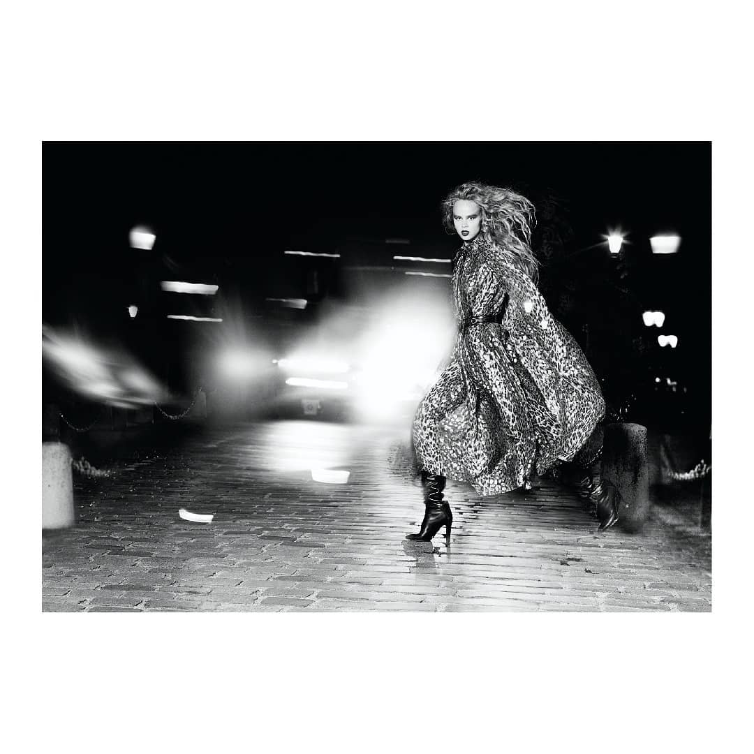A black and white fashion shot of a woman in a leopard print coat walking on a cobblestone street at night, with blurred car headlights behind her.