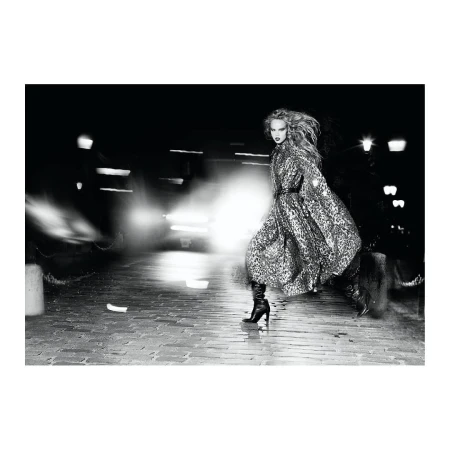 A black and white fashion shot of a woman in a leopard print coat walking on a cobblestone street at night, with blurred car headlights behind her.