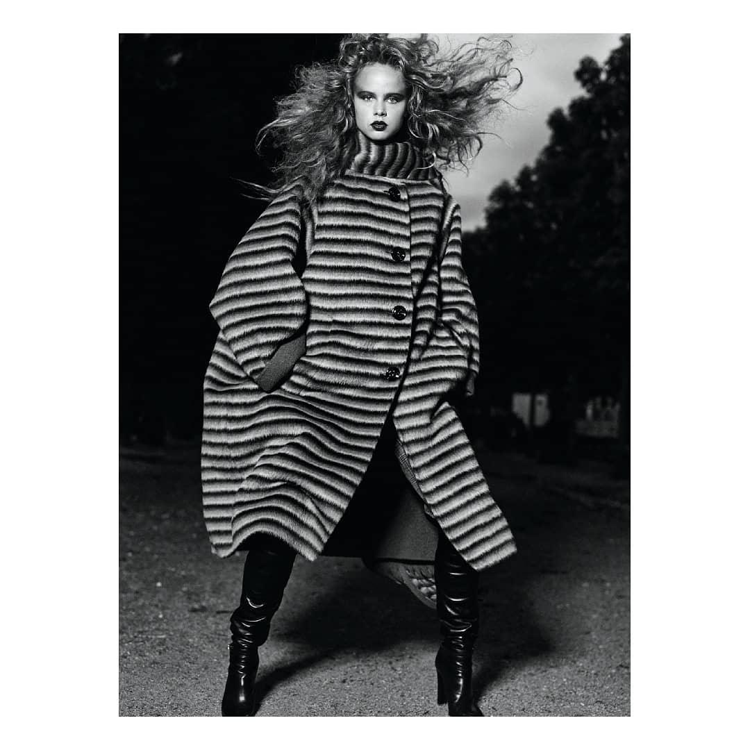 Black and white portrait of a woman with voluminous curly hair wearing a striped coat and tall boots outdoors