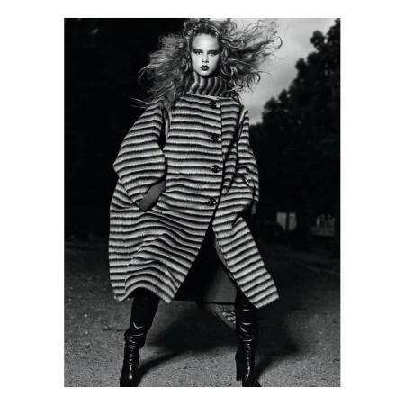 Black and white portrait of a woman with voluminous curly hair wearing a striped coat and tall boots outdoors