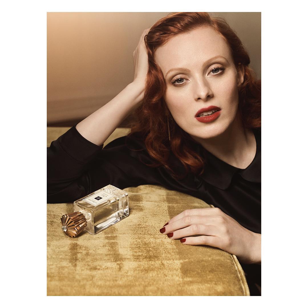 Red-haired woman poses with a perfume bottle on a velvet couch in a warm, inviting indoor setting, creating a sense of elegance.