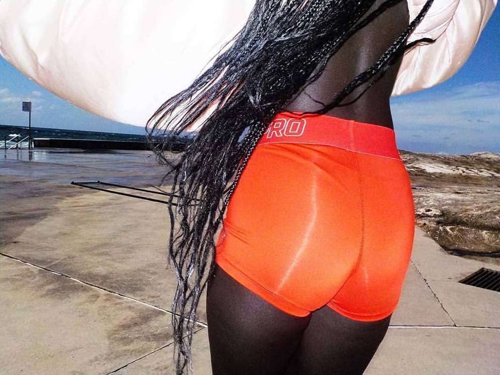 Athletic woman in orange compression shorts with black hair poses on a concrete platform by the ocean with an overcast sky.
