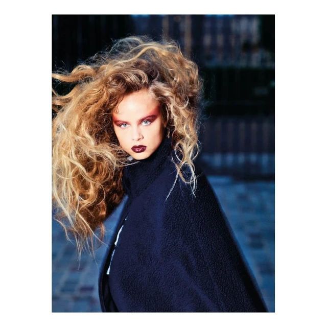 Woman with dramatic blonde curly hair and makeup, wearing a black cape, poses in front of a brick wall in an outdoor setting.