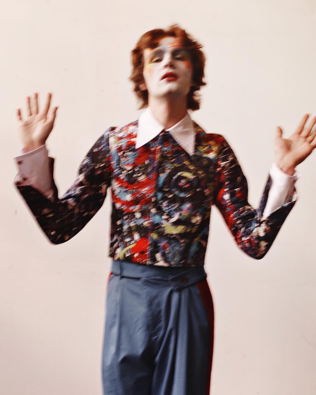 A person with heavy stage makeup and a patterned shirt raises their hands in a studio shot, creating a theatrical and artistic image.